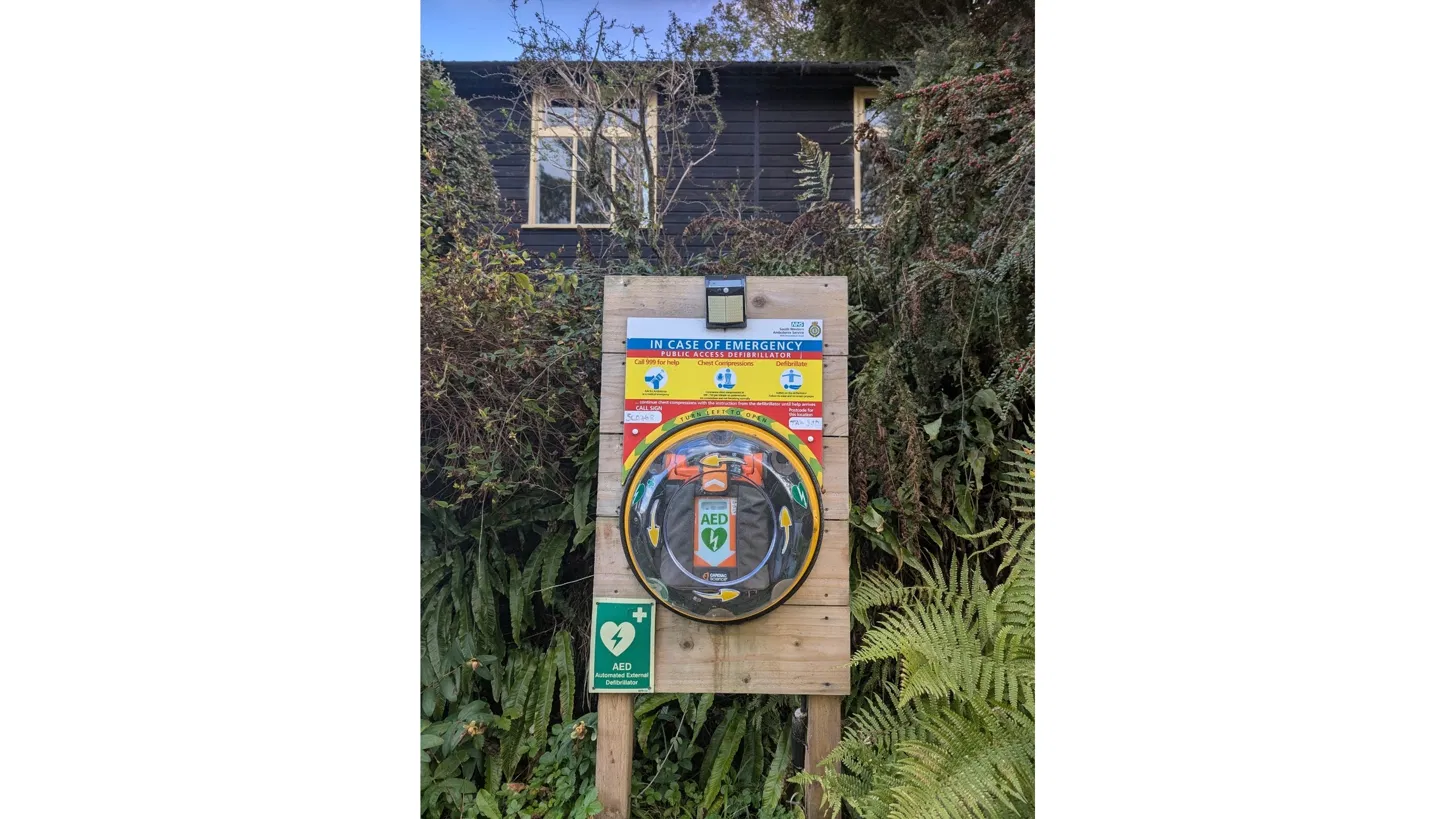 Public access defibrillator in a yellow cabinet on a wooden post outside Combe Florey Village Hall